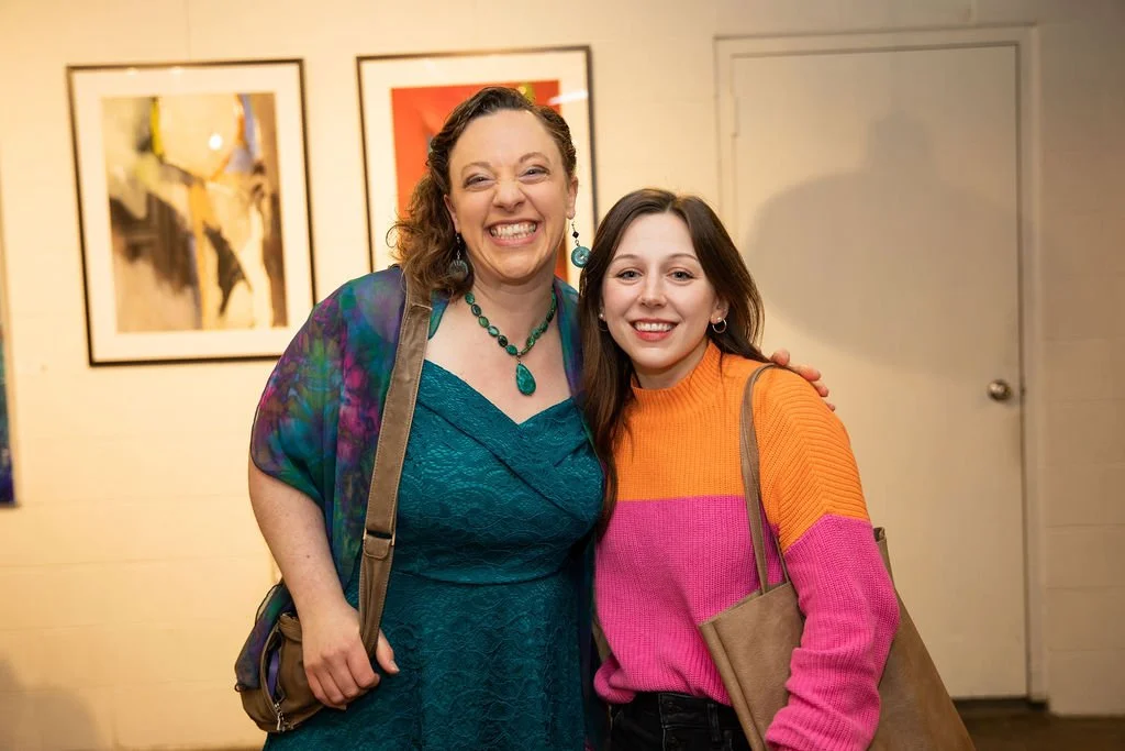 Two women smiling in an art gallery, woman on left wearing a teal dress and colorful shawl, woman on right in an orange and pink sweater. Framed artwork in the background.
