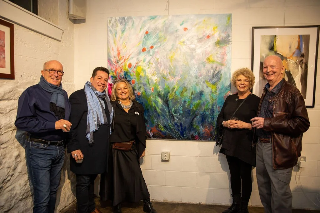 A group of five people standing in an art gallery, smiling, with vibrant abstract paintings on the walls.