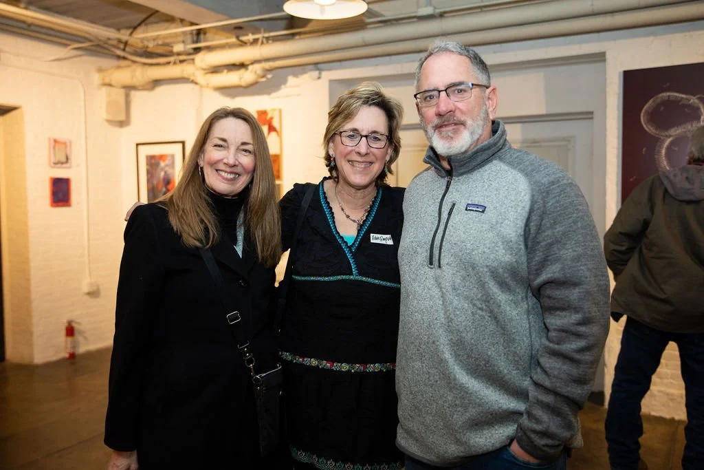 Three people posing and smiling indoors at an art gallery, with various artworks displayed on the walls behind them.