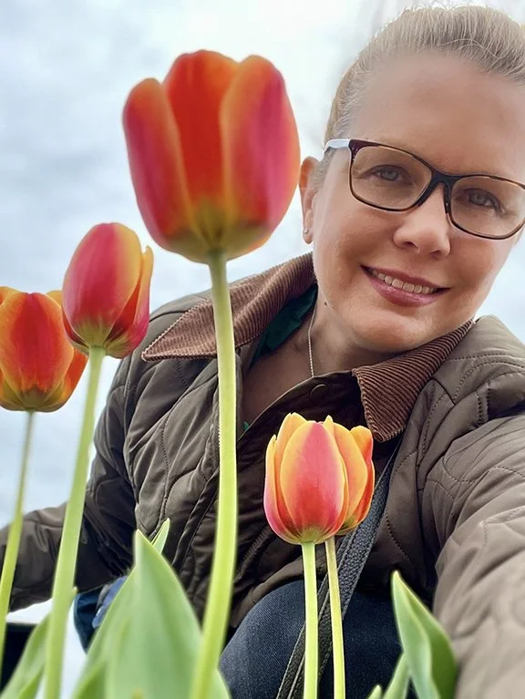 A woman with glasses taking a selfie among colorful orange and pink tulips outside on a cloudy day.