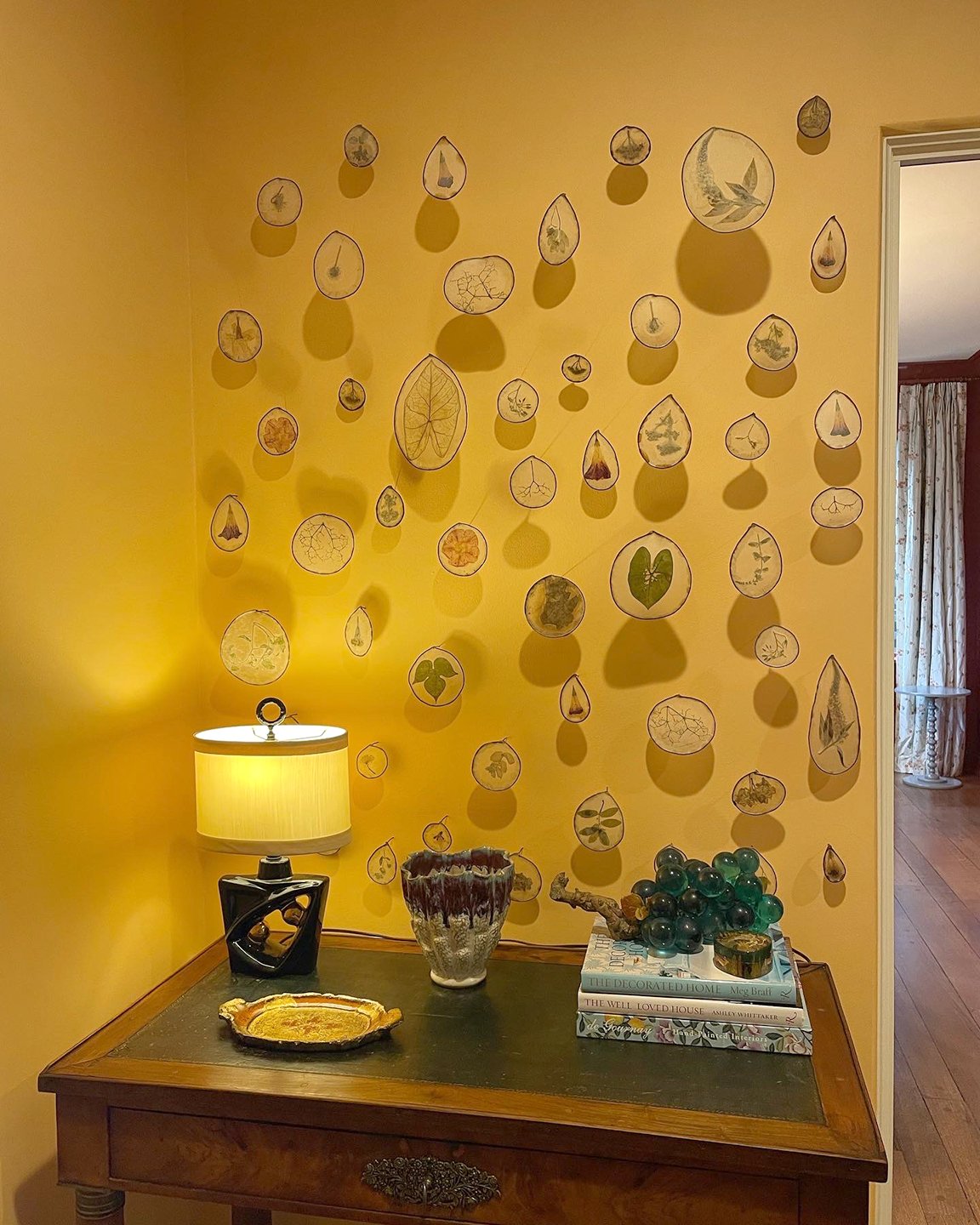 Yellow wall with framed pressed leaves and botanical illustrations hanging above a wooden table. The table has a table lamp, a decorative pottery vase, a stack of books, a glass grape sculpture, and a ceramic dish.