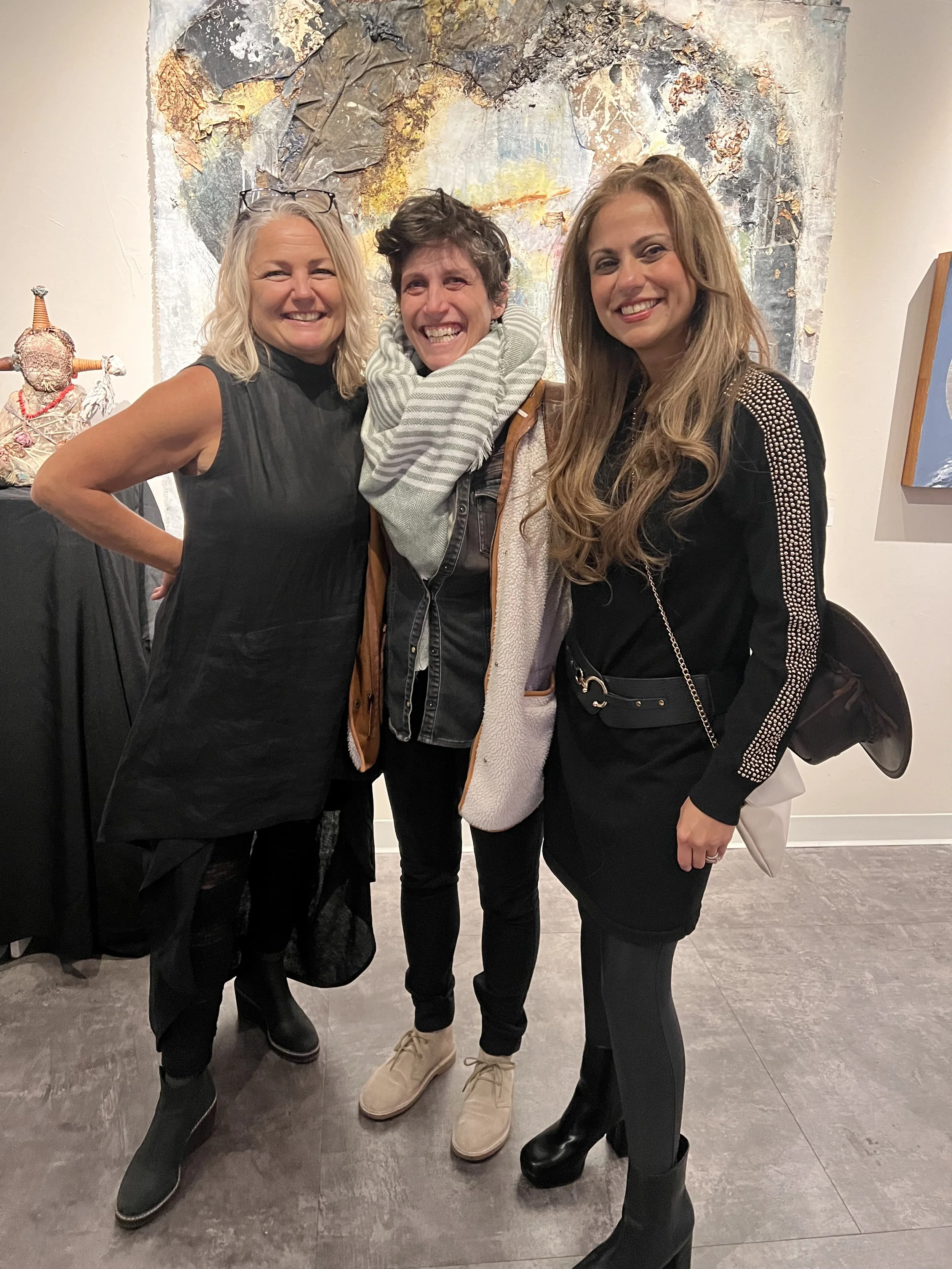 Three women standing together at an art gallery, smiling, with artwork behind them.