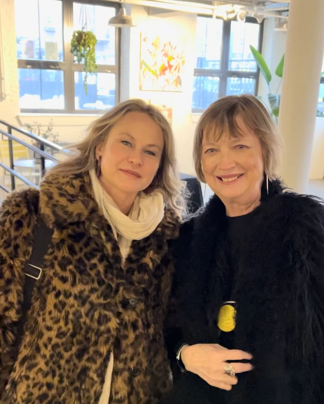 Two women in a brightly lit room, both wearing fur coats, one leopard print, smiling towards the camera. Art and plants are visible in the background.