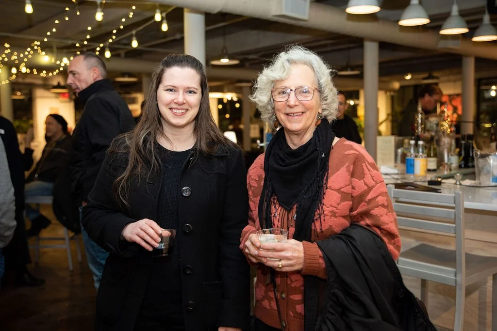 Two people smiling and holding drinks at a social event in a warmly lit indoor setting with a bar in the background.
