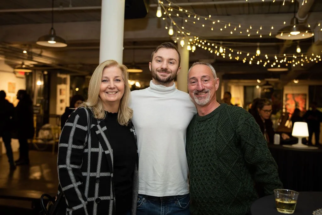 Three people posing together in a warmly lit event space with string lights. The person on the left is wearing a black and white checkered jacket, the person in the middle a white turtleneck, and the person on the right a green knitted sweater. Peopl