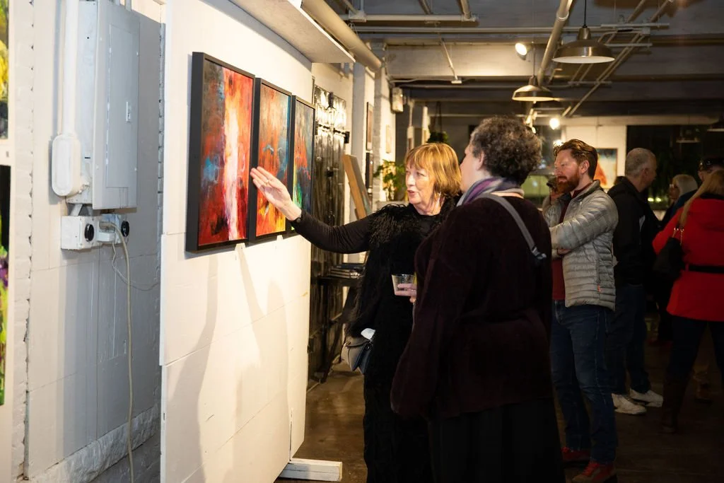Art gallery with people viewing abstract paintings, one woman pointing at a colorful artwork on the wall.