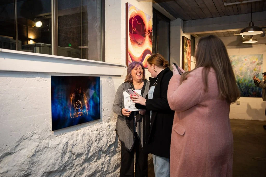 A group of three people in an art gallery. They are standing together, smiling and interacting. Various colorful abstract artworks are displayed on the walls around them.