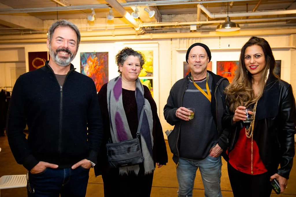 A group of four people posing in an art gallery. The setting is indoors with art on the walls. They are casually dressed and each holding a drink. The lighting is warm and the atmosphere appears relaxed and social.