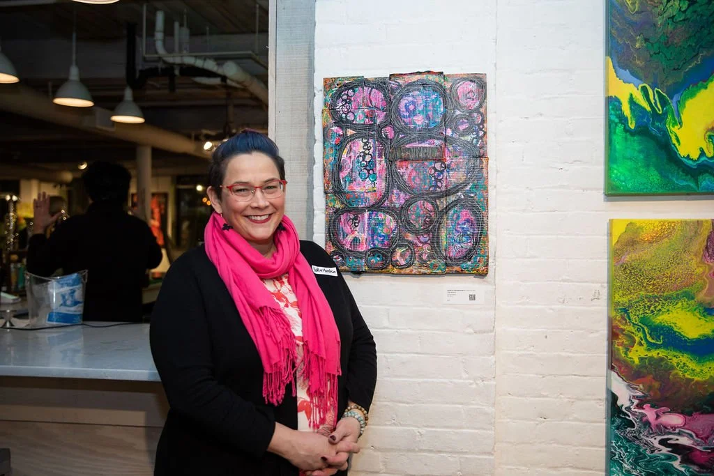 Woman in a black jacket and pink scarf standing next to abstract artwork on a white brick wall in an art gallery.