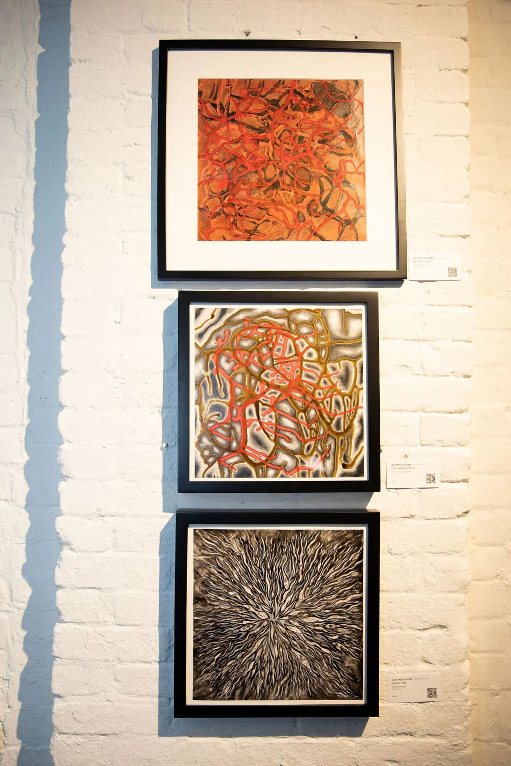 Three framed abstract artworks displayed on a white brick wall. The top piece features orange and black swirling patterns. The middle artwork has a mix of red, brown, and gray, creating a chaotic web-like design. The bottom artwork shows a radial bur