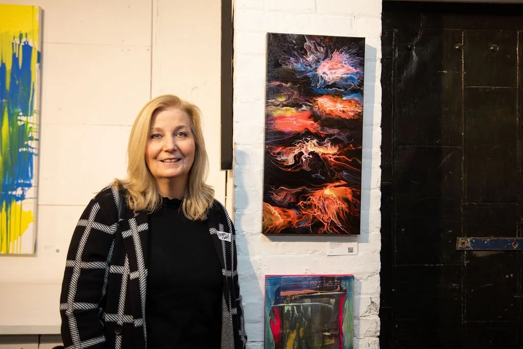 A woman standing next to an abstract painting displayed on a white brick wall in an art gallery.