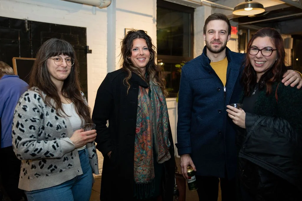 Four people at a casual indoor gathering, smiling and holding drinks, with a brick wall and windows in the background.