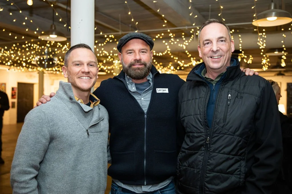 Three men posing together at an indoor event with string lights in the background.