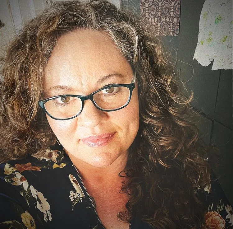 A woman with curly brown hair and glasses smiling at the camera.