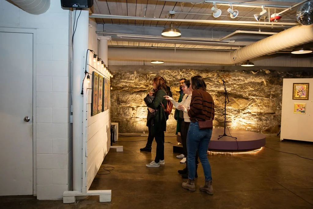 People viewing artwork at a gallery exhibition with a rock wall background and small stage with a microphone.