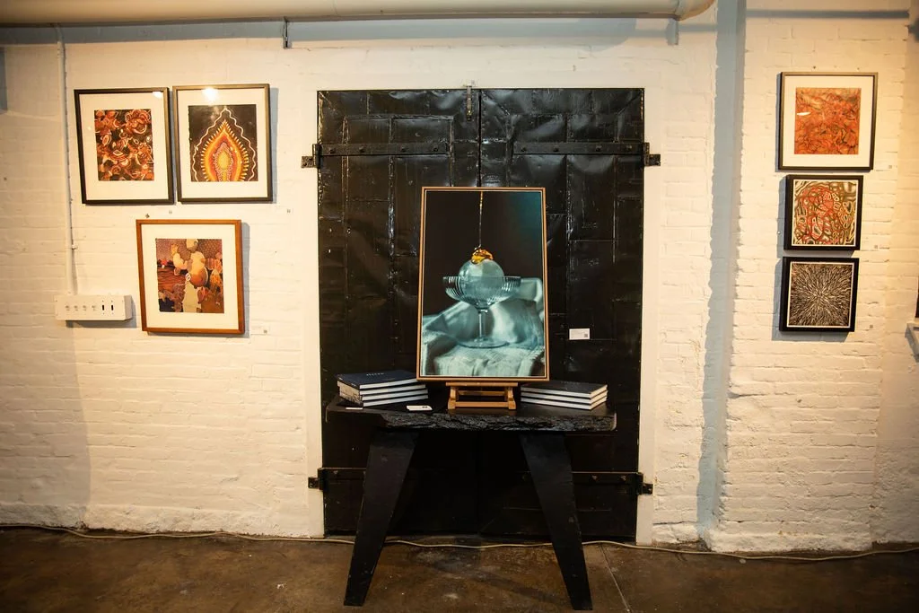 Art gallery wall with various framed artworks, including abstract paintings and a photograph of glassware. A black table holds additional seating or decorative objects.