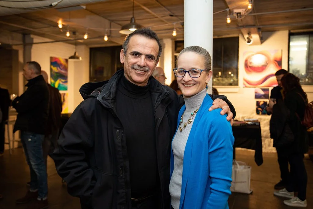Two people smiling at a gallery event, surrounded by artwork and other attendees.