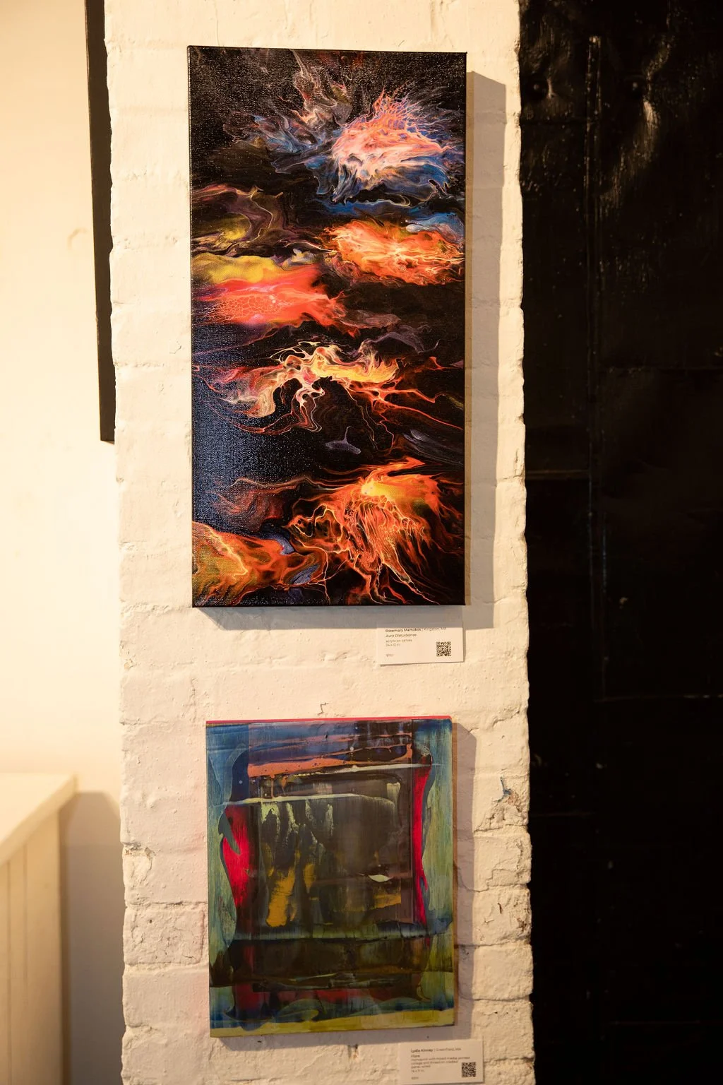 Two abstract paintings on a white wall, one with vibrant swirling colors and the other with darker, layered hues.