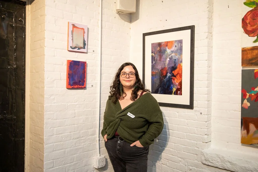 A person with glasses and long hair, wearing a green sweater, stands in an art gallery. They are surrounded by abstract paintings on a white brick wall. The artwork features bold, colorful designs, with one framed piece prominently displayed beside t