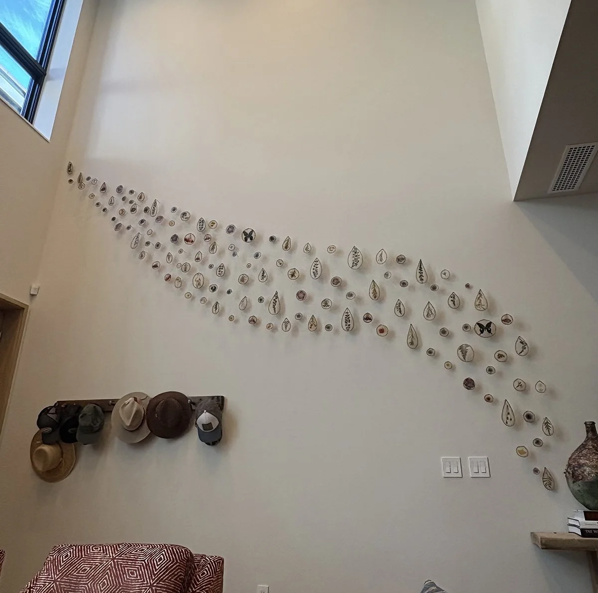 Decorative wall art featuring small, leaf and nature-themed illustrations arranged in a curved pattern on a white wall, with hats hanging on a wooden rack and a corner vase with greenery at the bottom right.