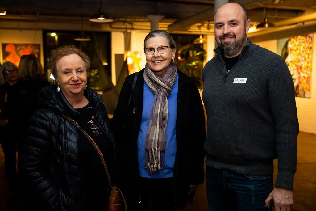 Three people smiling at an indoor art gallery event, with paintings visible in the background.