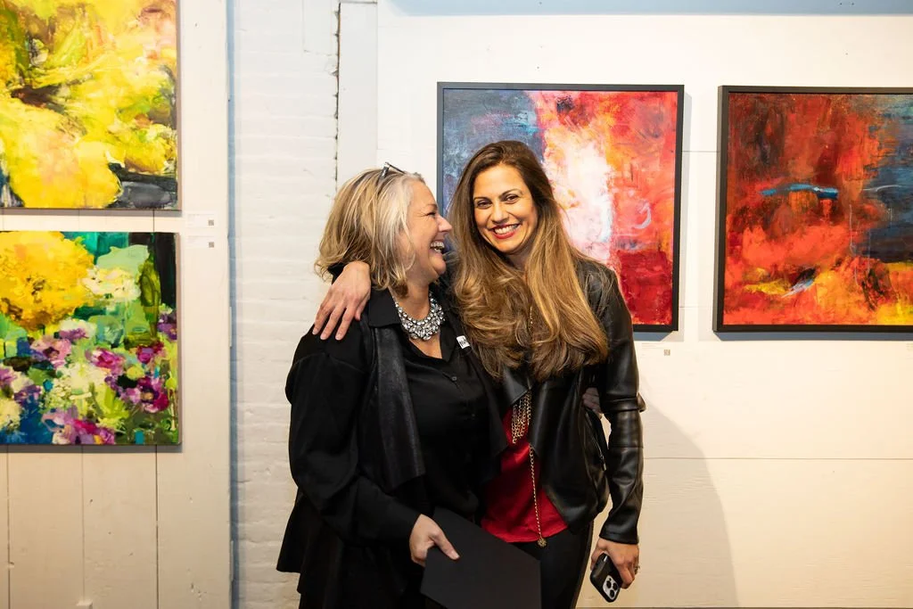 Two women smiling in an art gallery with colorful abstract paintings on the wall behind them.