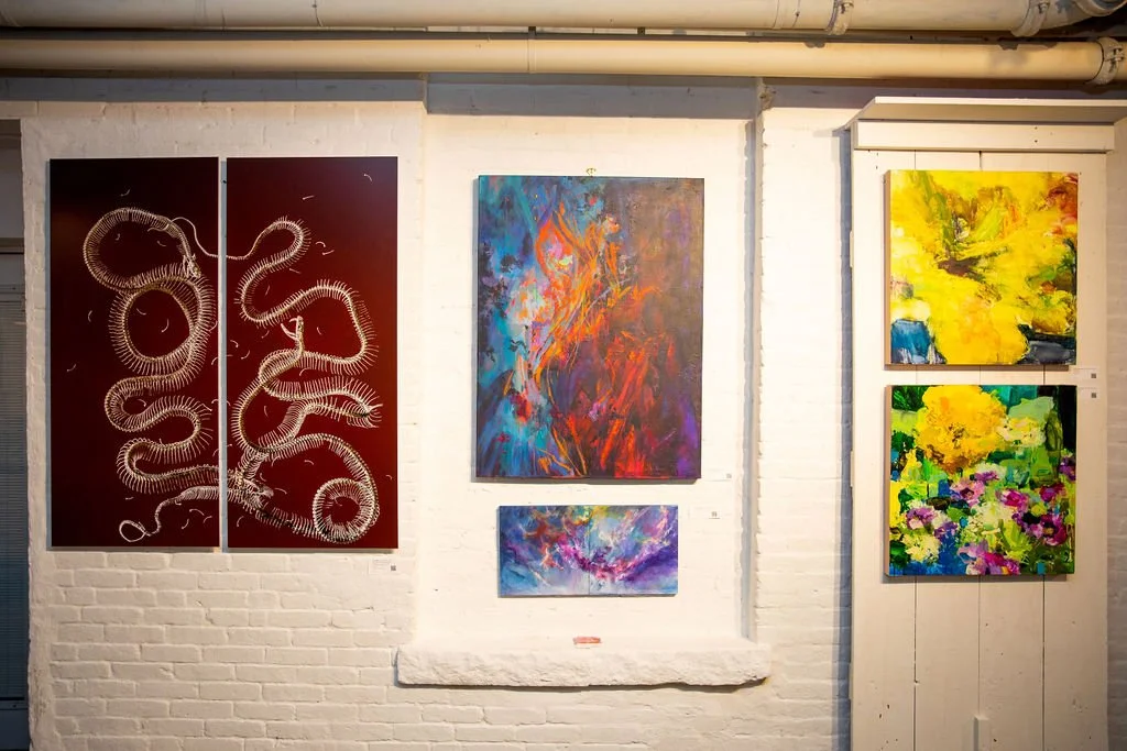 Gallery wall with five abstract paintings, featuring diverse styles and colors, including reds, blues, yellows, and greens, on a white brick background.