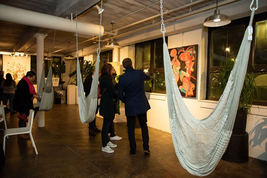 Art gallery interior with people viewing paintings, hanging hammocks, and potted plants.