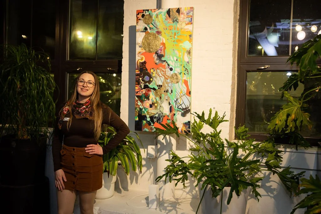 Woman standing next to colorful abstract painting in a room with large plants.