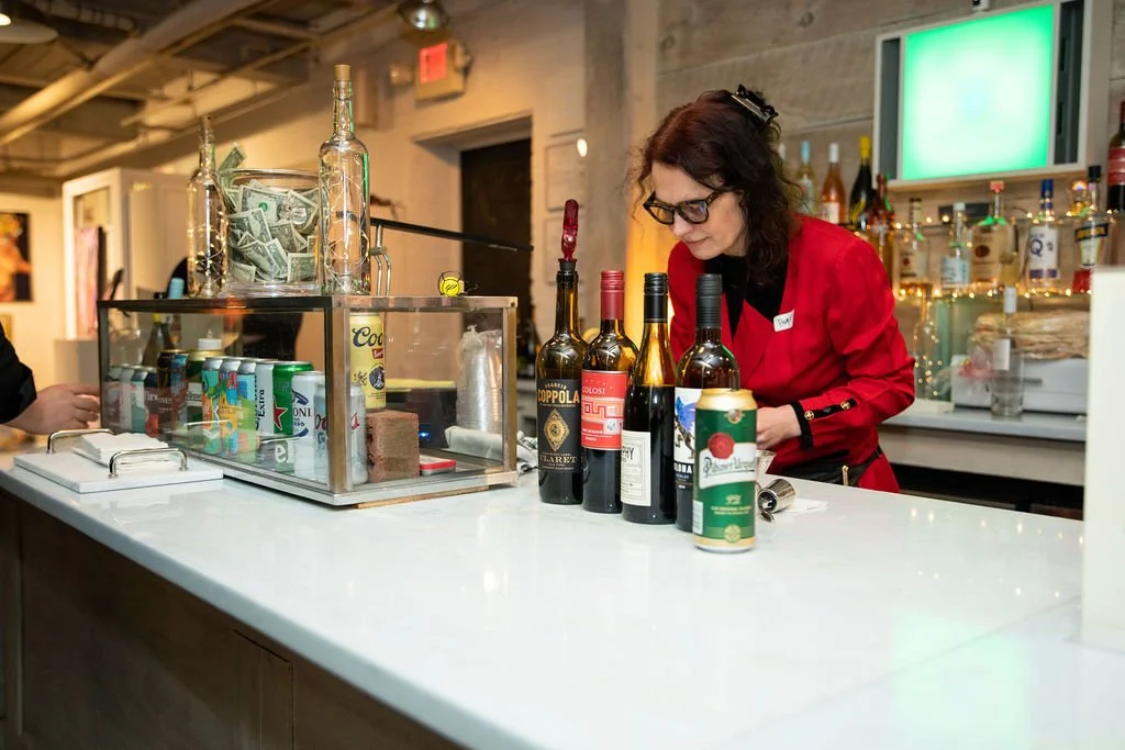 Bartender in red serves drinks at a bar with wine bottles and canned beverages displayed.