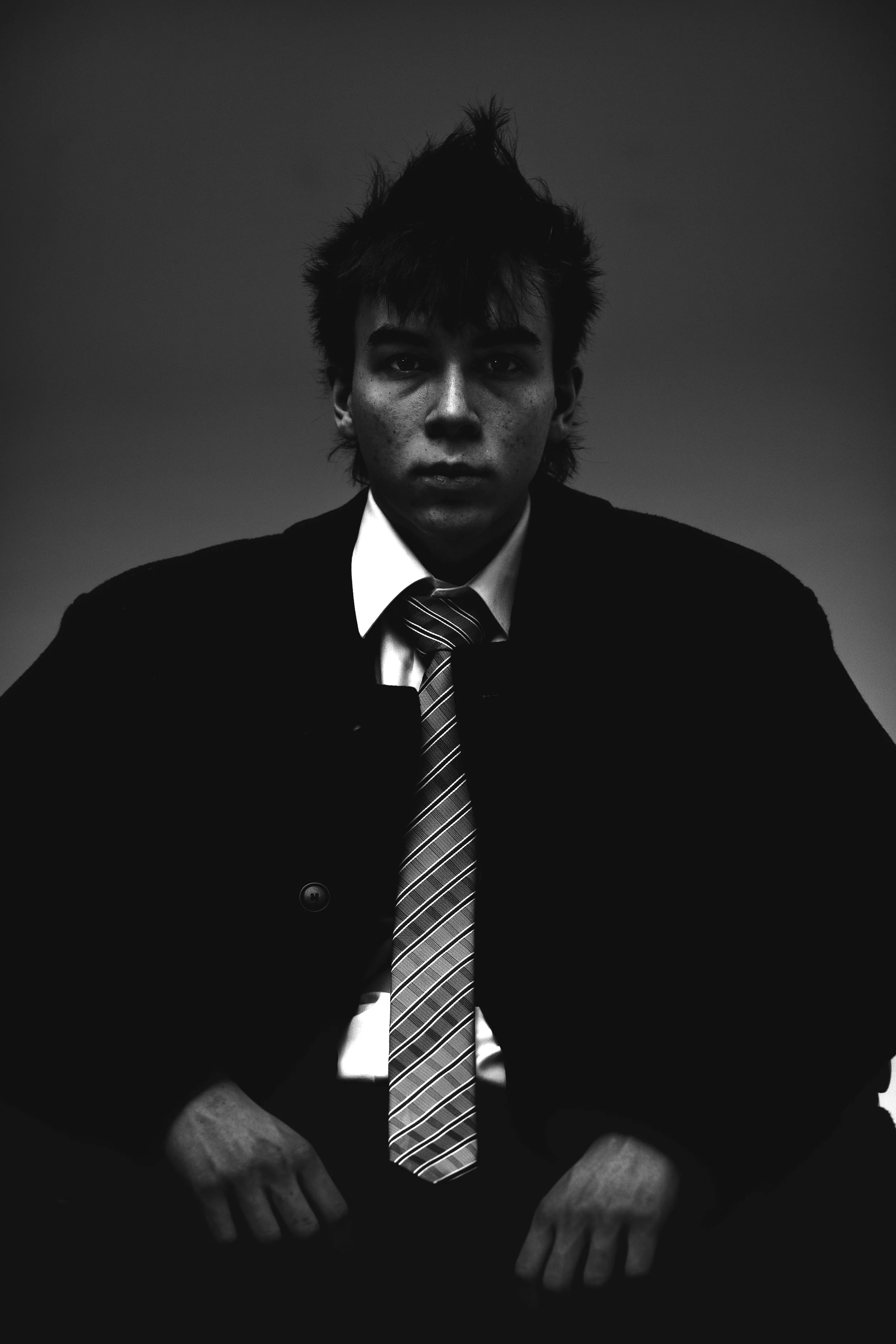 Black and white photo of a young man with a serious expression, messy hair, dressed in a suit and tie, looking directly at the camera.