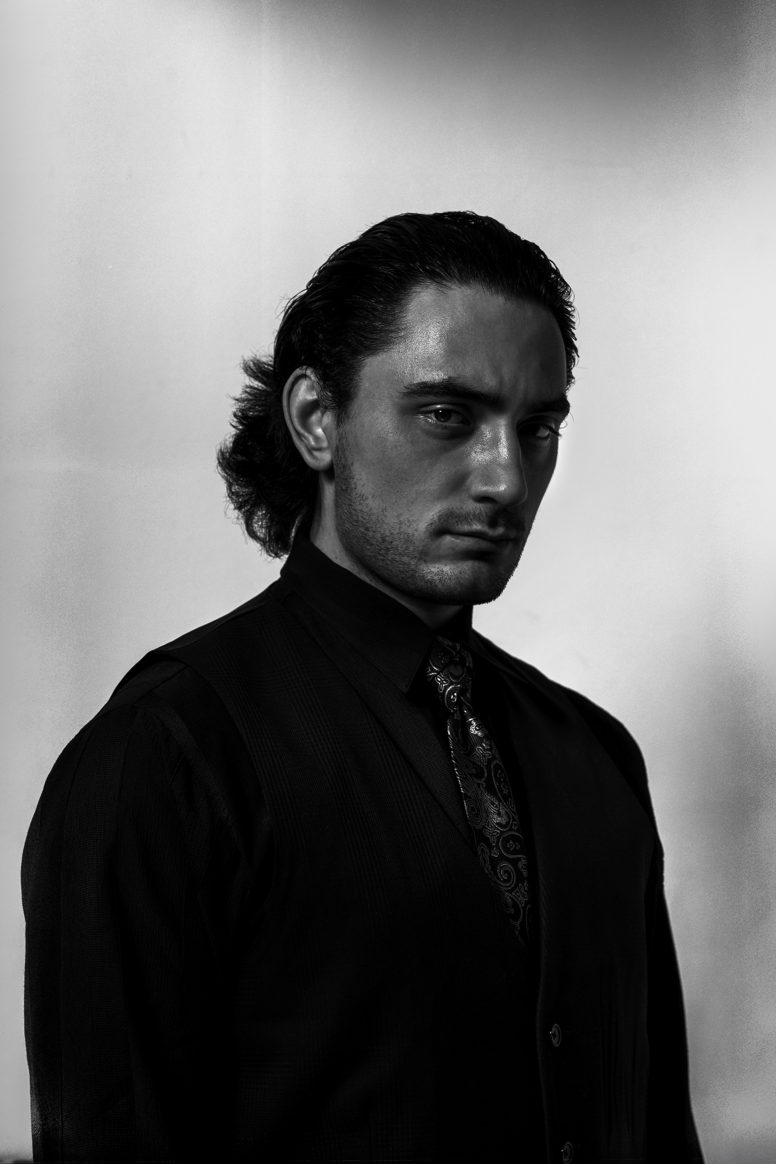 Black and white portrait of a man with slicked-back hair, wearing a dark shirt and patterned tie, gazing intensely at the camera.