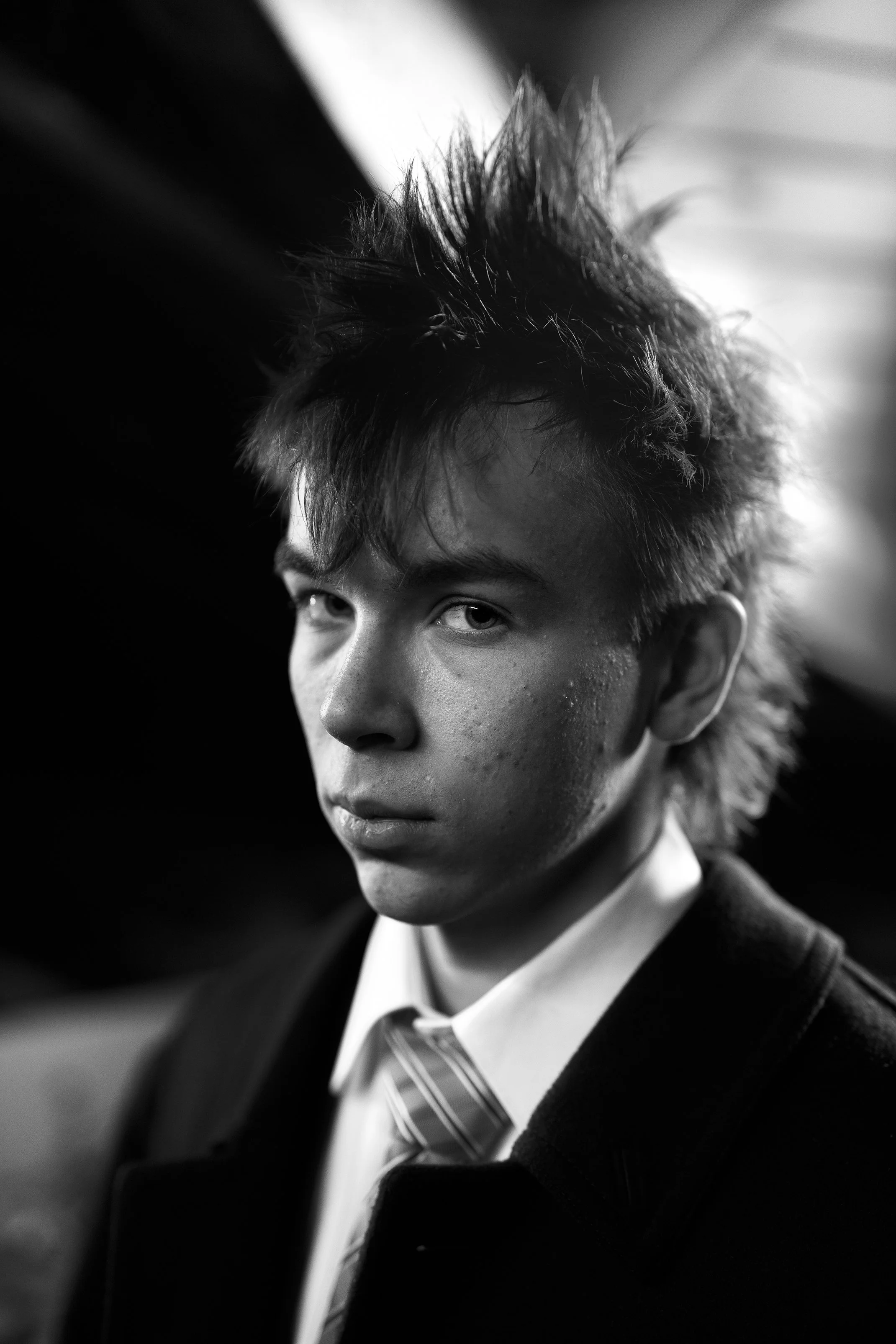Black and white portrait of a young man with spiky, messy hair wearing a suit and tie, looking intensely at the camera.