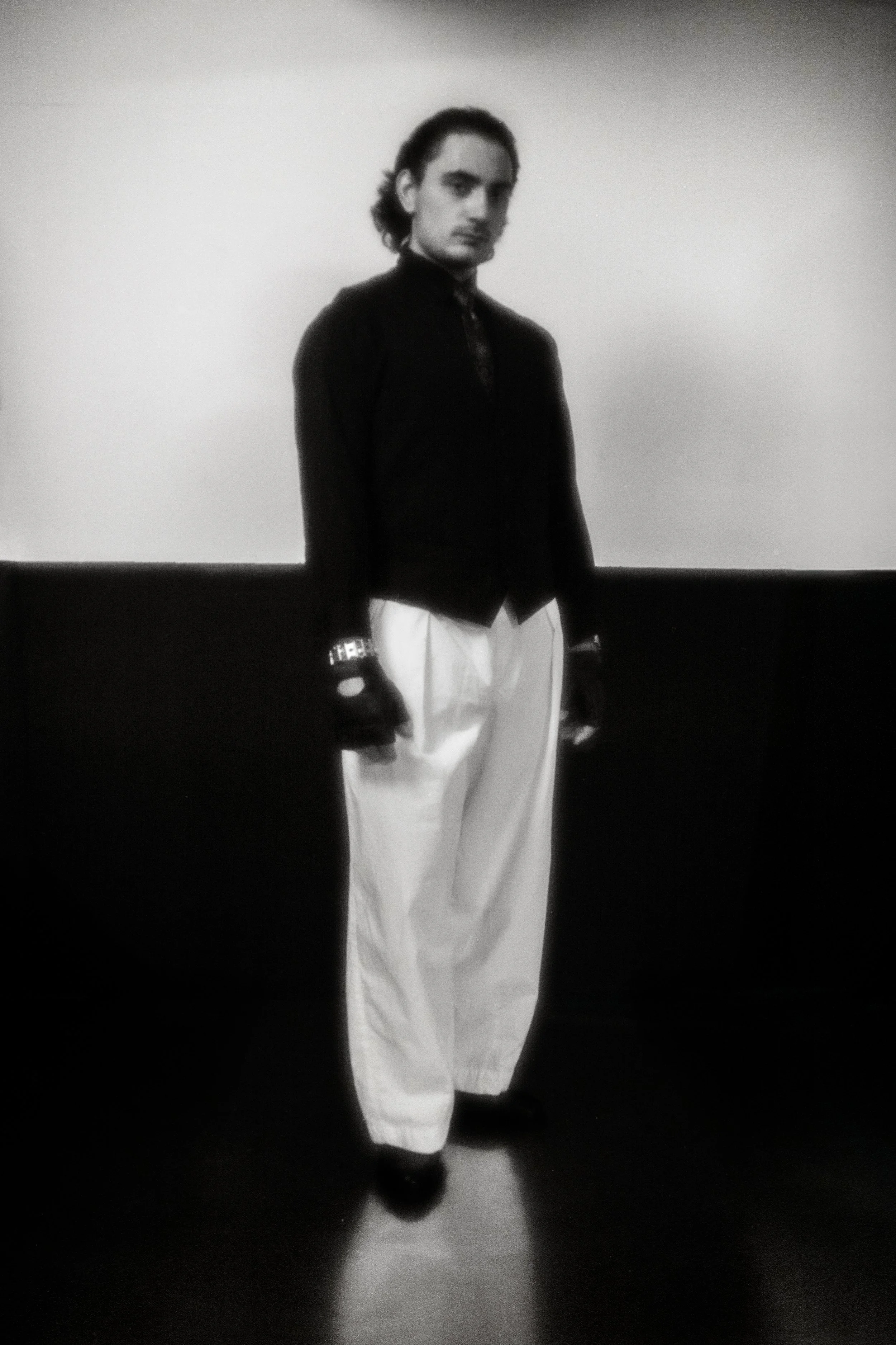 Black and white photo of a young man standing against a background, wearing a dark jacket with gloves and loose white pants.