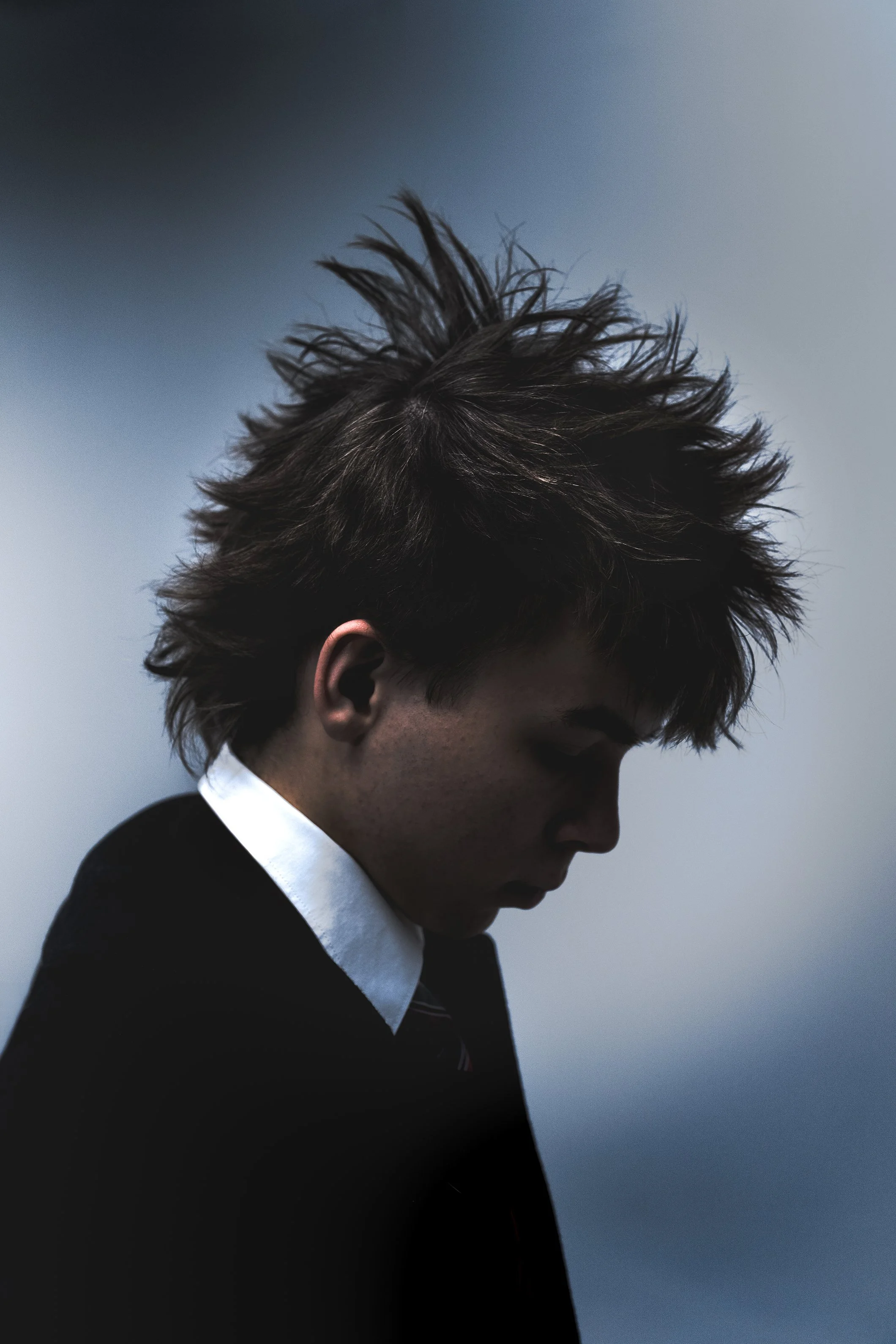 A young man with spiky, tousled hair, wearing a black jacket and white collared shirt, looking down against a blue-gray background.