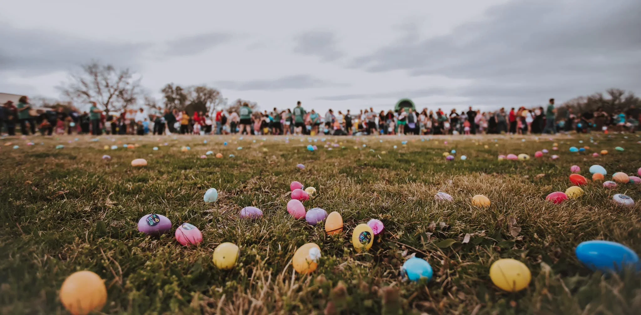 A large group of people gathered outdoors on a cloudy day, with colorful plastic Easter eggs scattered across the grass in the foreground.