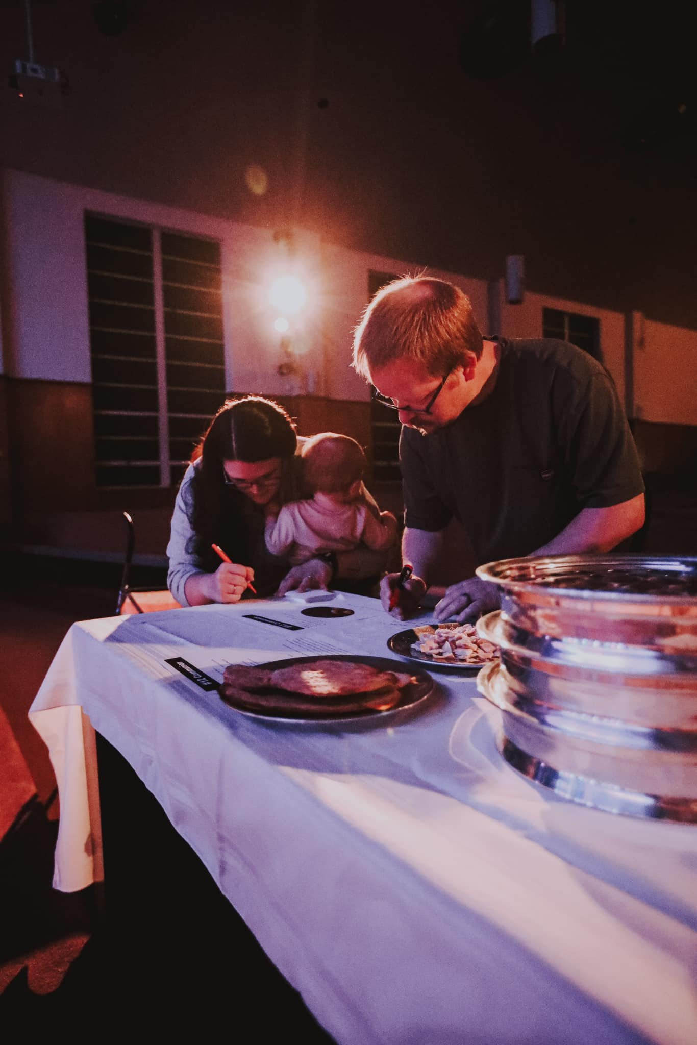 People gathered at a table, with food and trays, in a dimly lit room, possibly during an event or dinner.