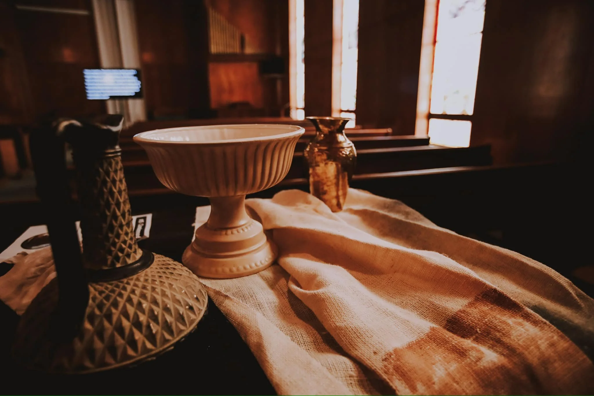 a close-up of a decorative display with various vintage glassware and a ceramic bowl, set on fabric, inside a rustic church with wooden pews and stained glass windows in the background