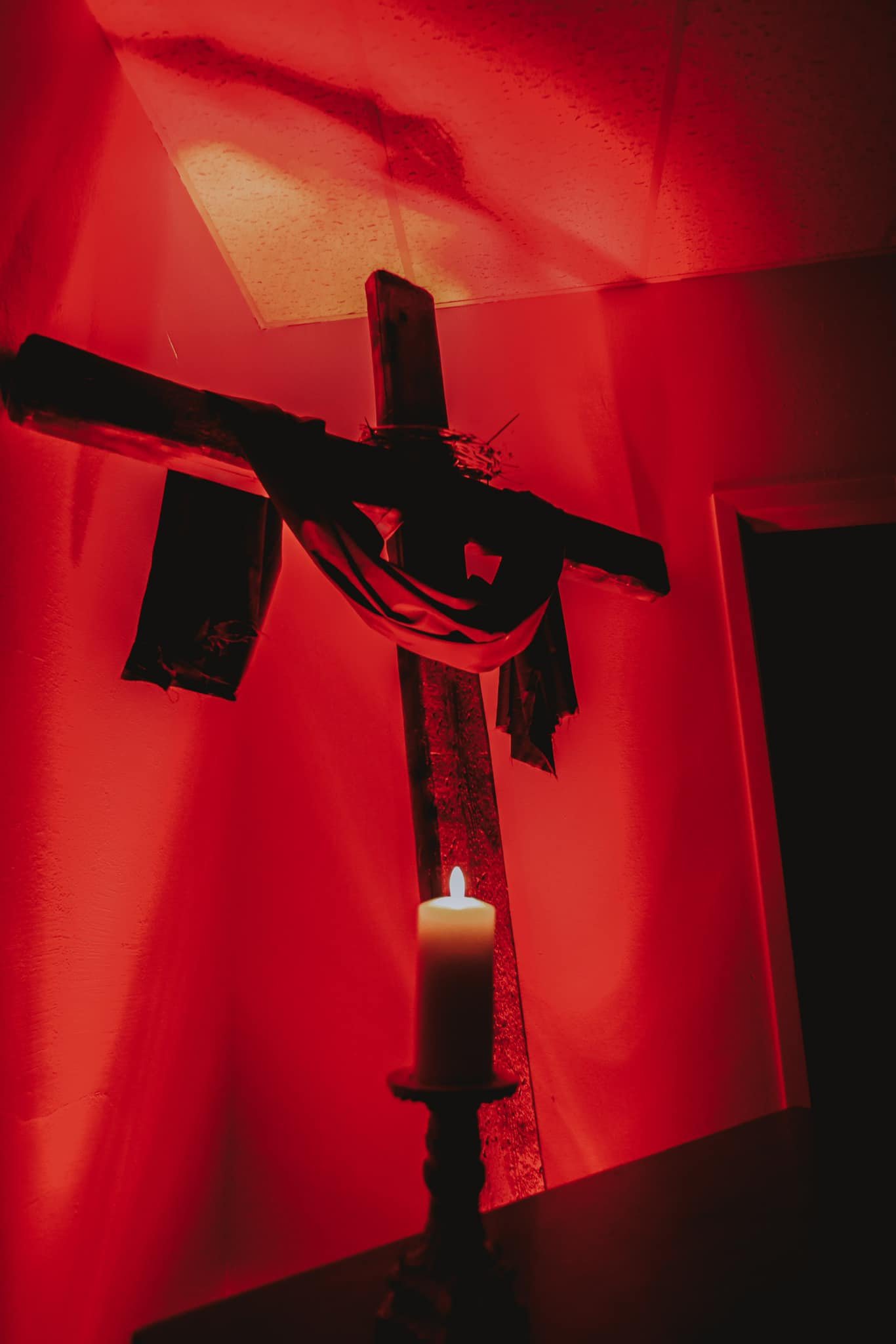 A wooden cross decorated with cloth hangs on a red wall. A lit candle is in the foreground.
