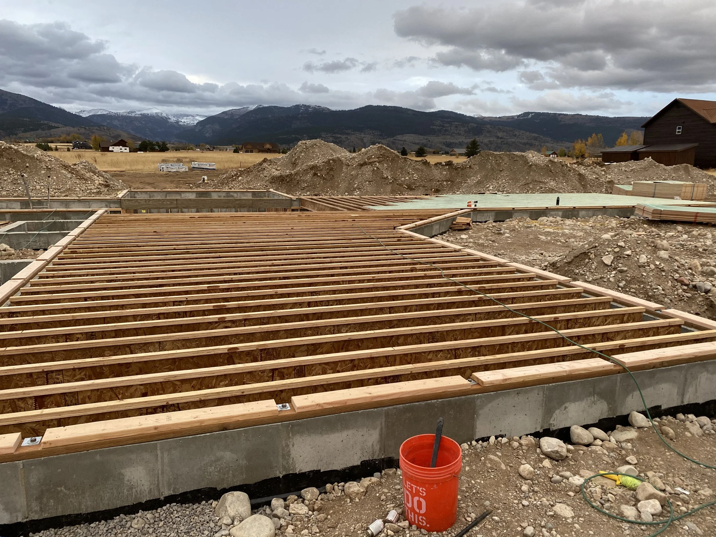 Construction site with wooden framing on concrete foundation, dirt mounds, and mountains in the background, overcast sky.