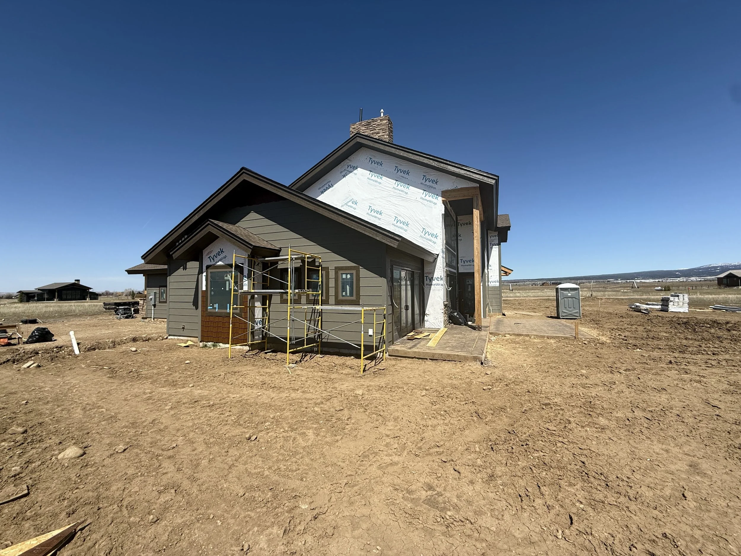 A house under construction with partially completed exterior walls and scaffolding in front. Construction tools and materials are scattered on the bare dirt ground surrounding the house under a clear blue sky.