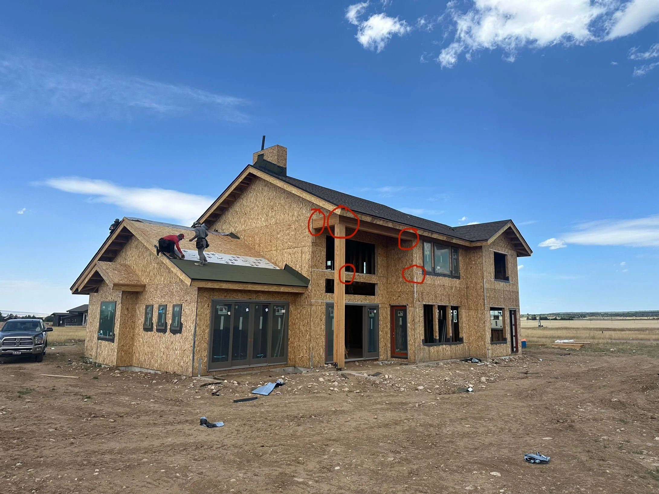 A house under construction with partially installed siding and a workers on the roof installing new shingles, on a dirt lot with a vehicle nearby and a clear sky overhead.