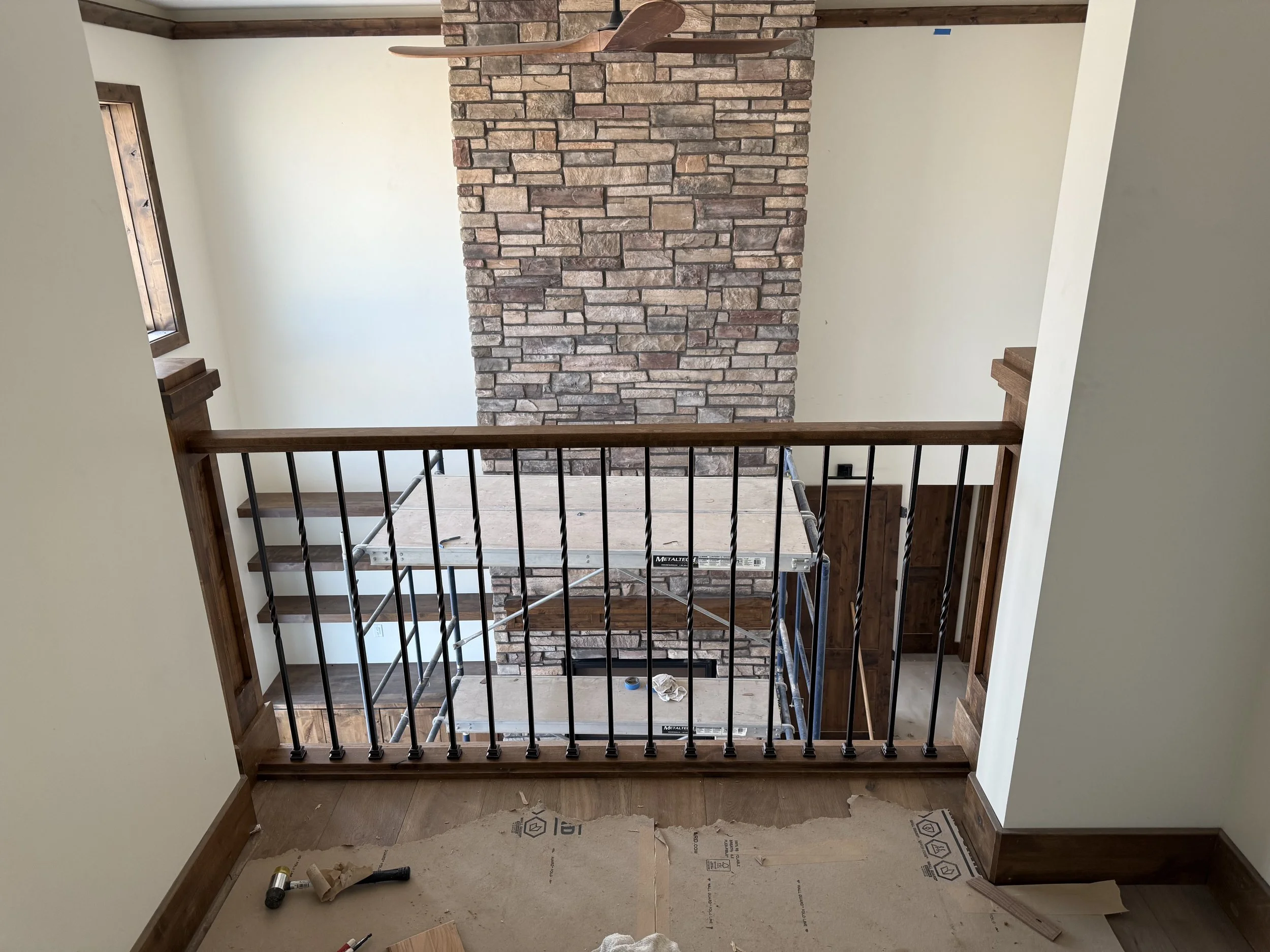 Interior view of a second-floor balcony with a black metal railing, wooden trim, and a brick fireplace visible in the background under construction.
