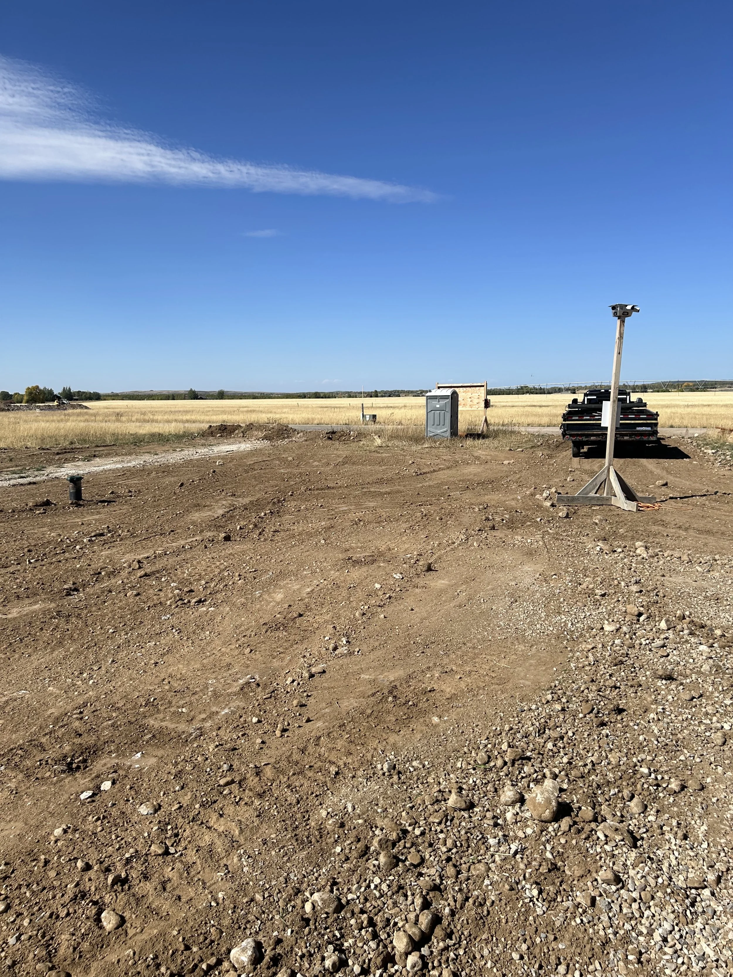 Construction site with dirt and gravel ground, a portable toilet, a parked pickup truck, and a tilted light pole against a clear blue sky.