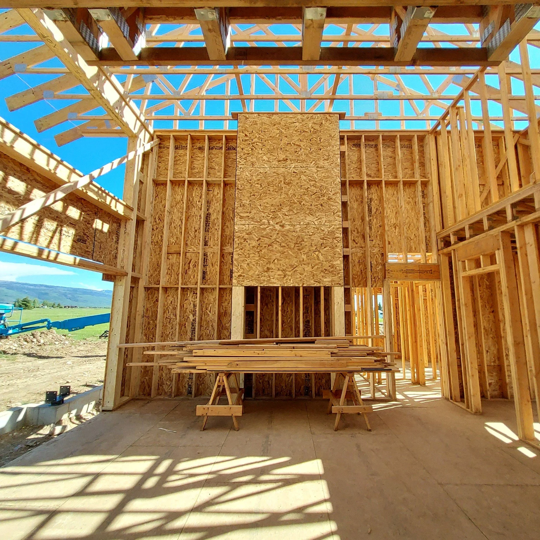 Interior view of a house under construction showing wooden framing and support beams, with some plywood panels installed on the walls. There is construction material stacked on tables in the center and sunlight streams through the open roof and frami