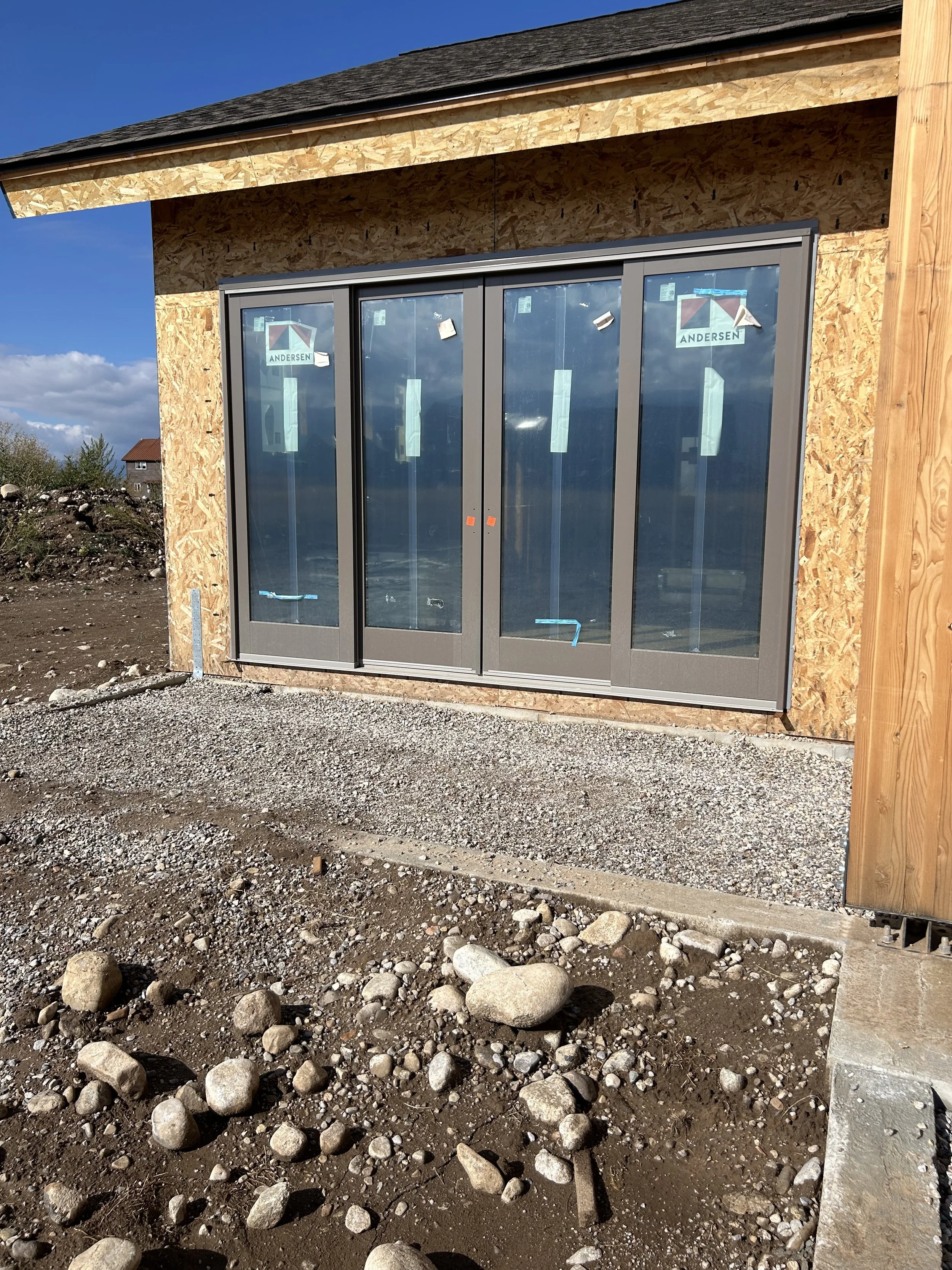 Construction site of a building with large glass sliding doors, framed in gray metal, in progress. The exterior wall has unfinished plywood sheathing, and the ground is covered with dirt and rocks. The sky is blue with some clouds.