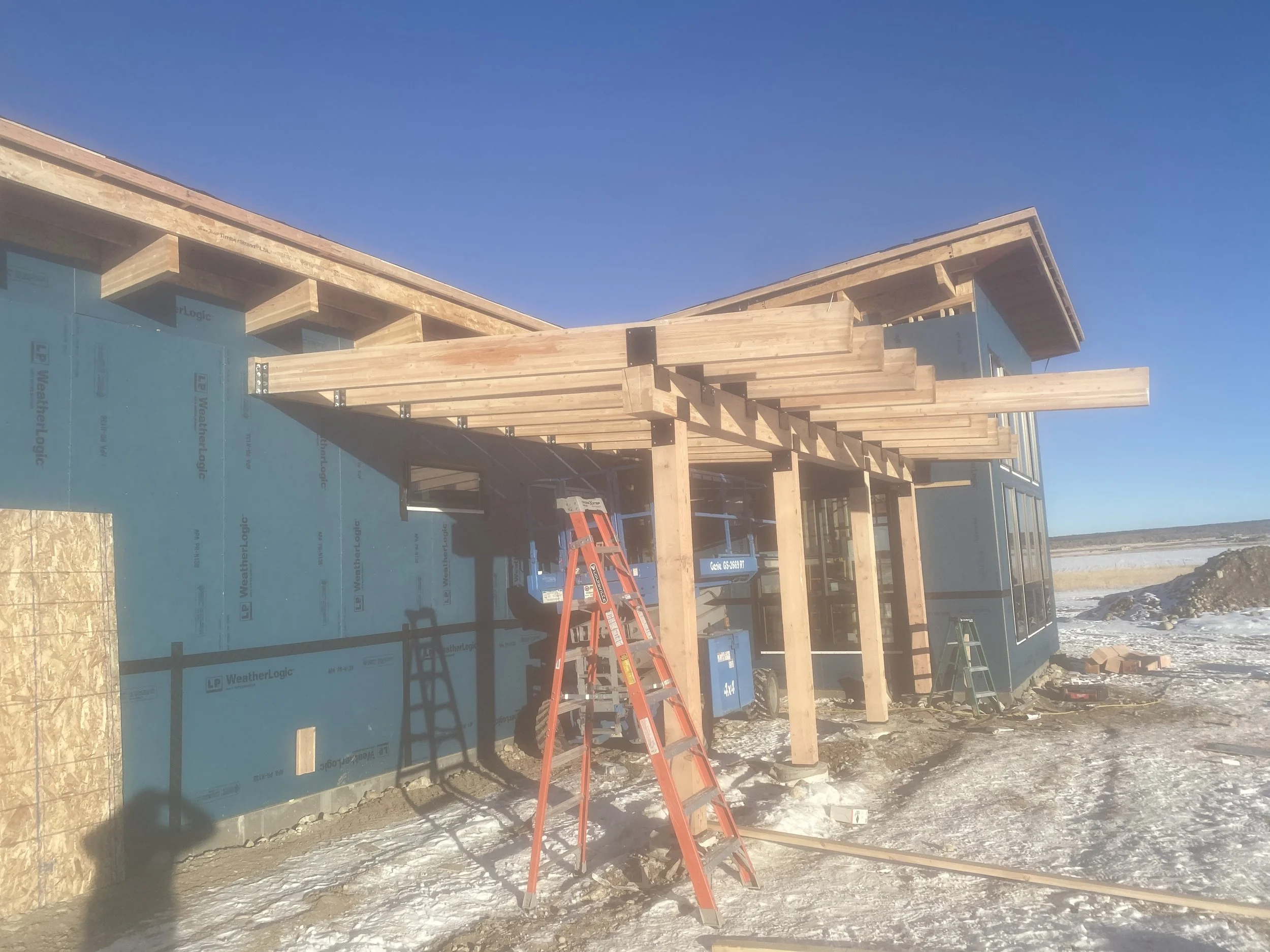 Construction site of a modern house with exposed wooden beams, blue weather-resistant siding, and a large glass window, set in a snowy landscape against a clear blue sky.