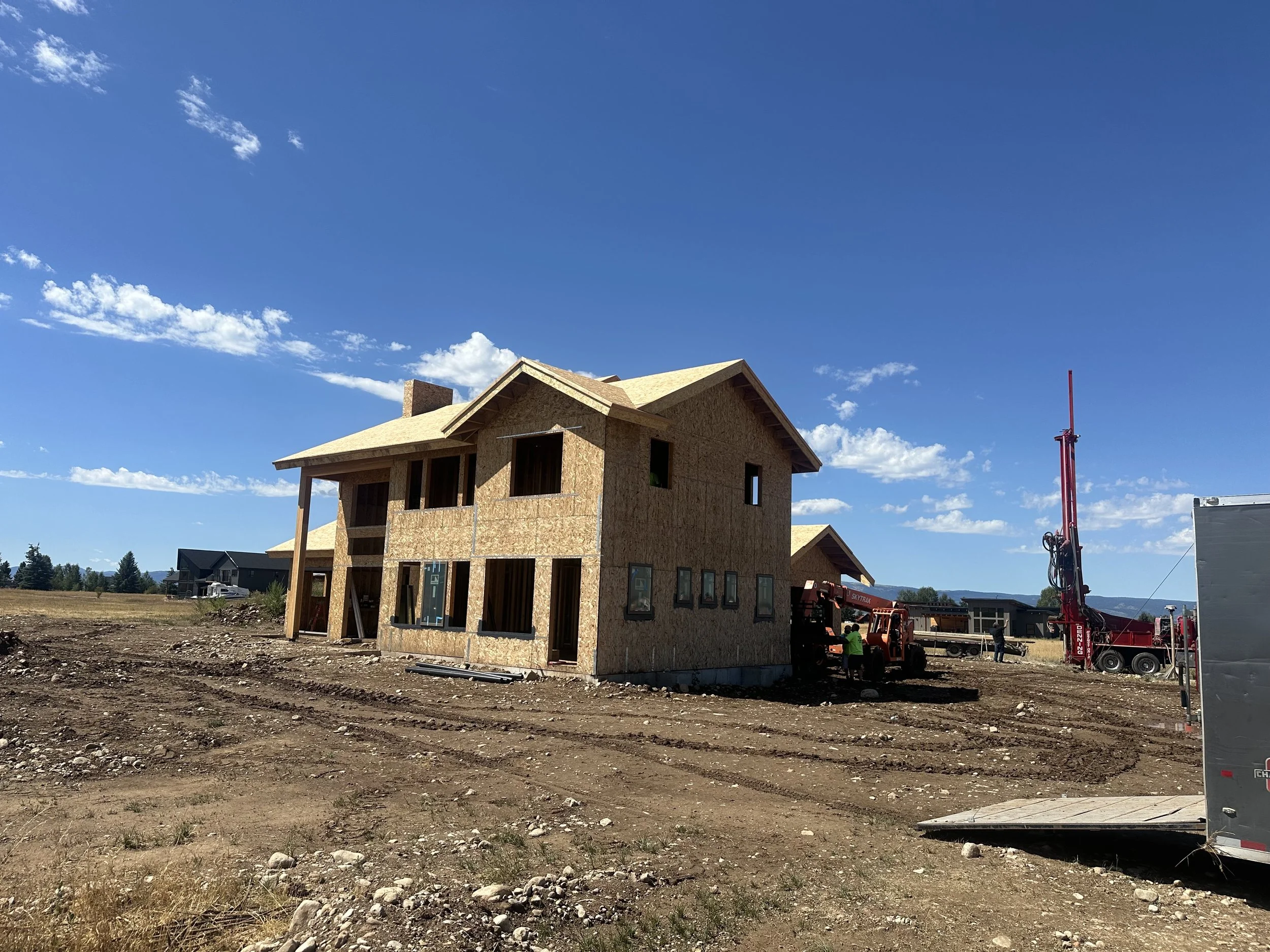 A two-story house under construction with wooden framing, surrounded by construction equipment, on a dirt lot with a clear blue sky.