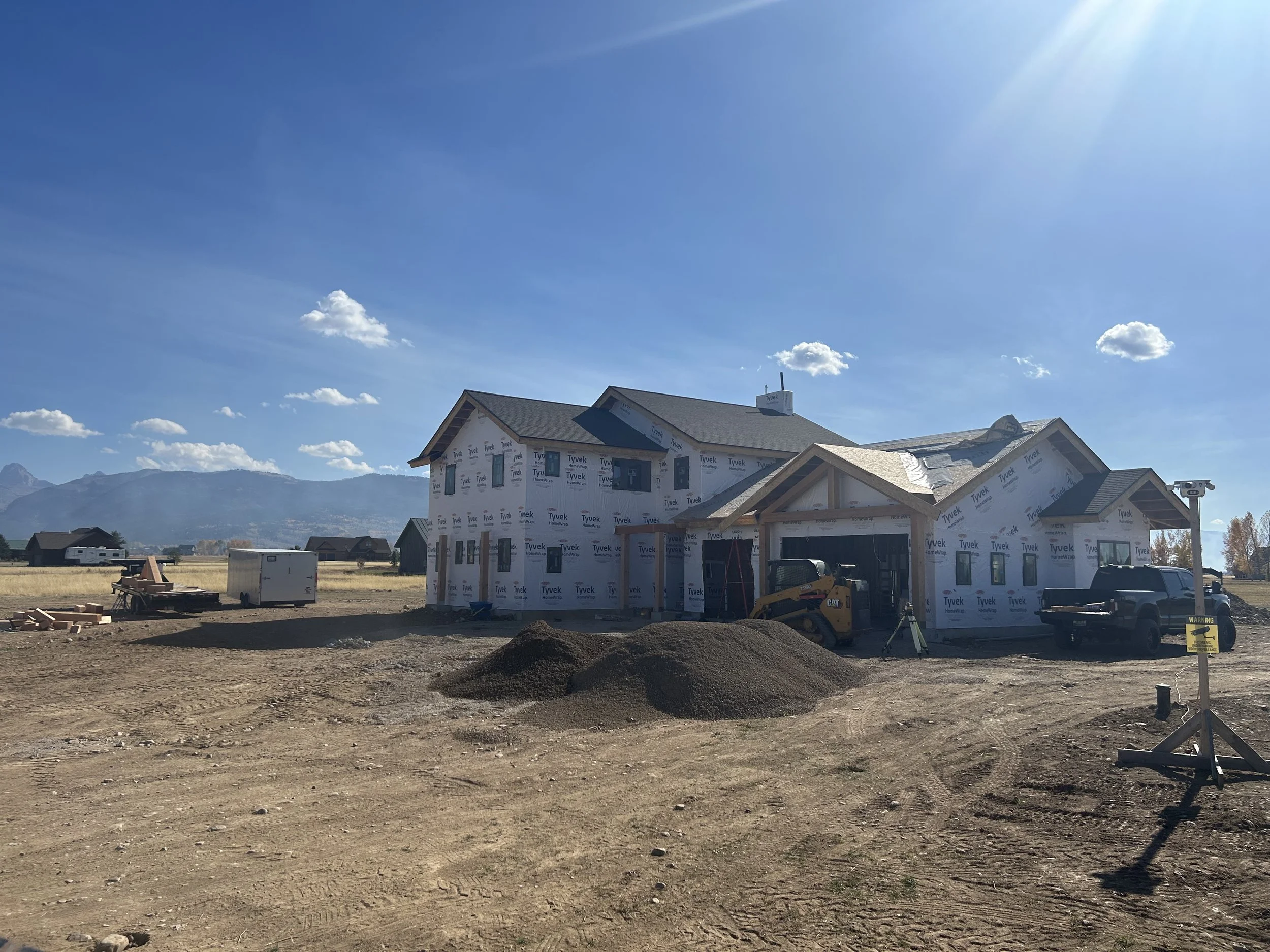 A house under construction with wrapped exterior walls, construction equipment, dirt piles, and a clear blue sky with mountains in the background.