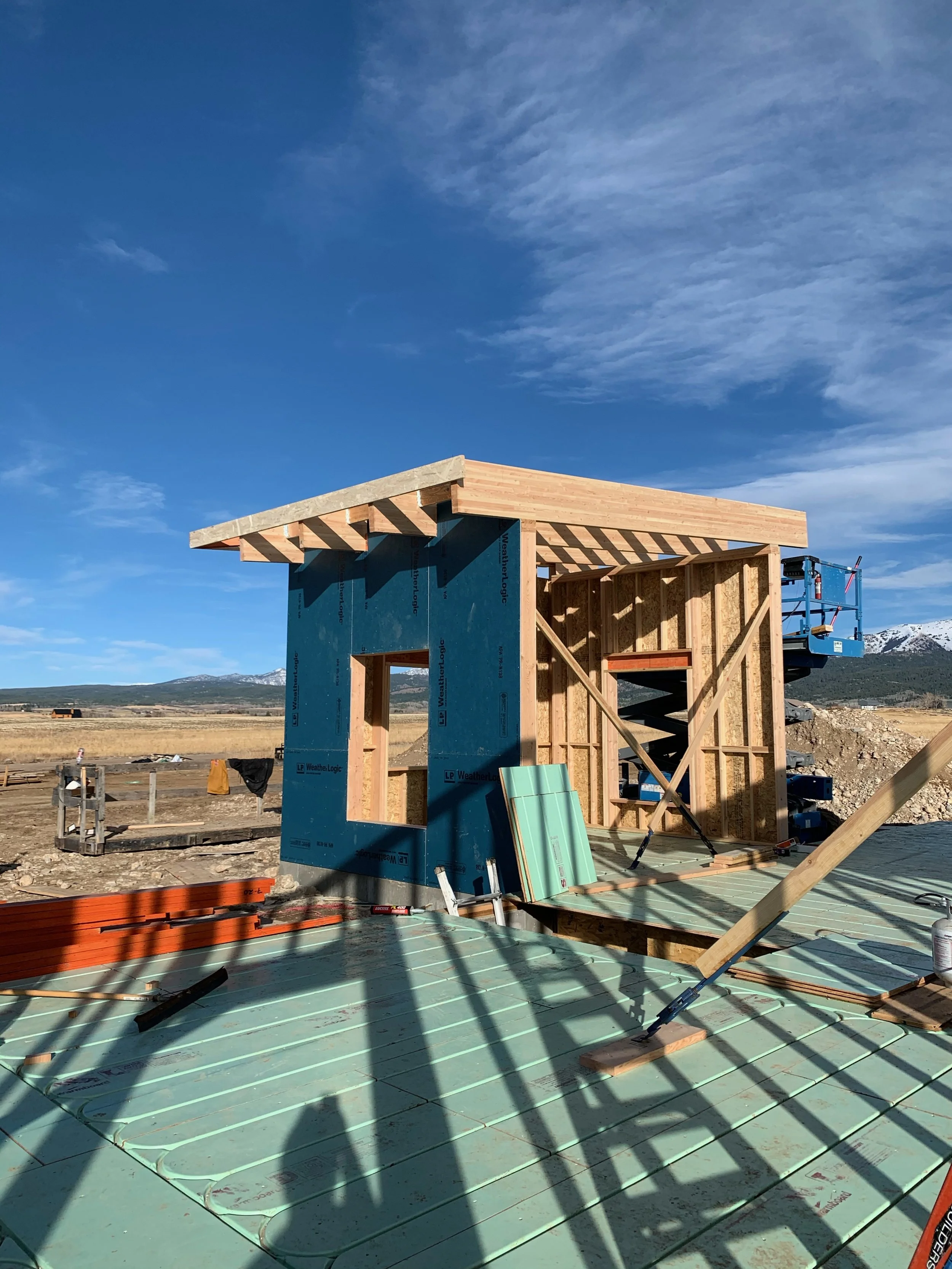 Construction site of a small wooden building with a sloped roof, blue weather-resistant sheathing on the walls, and green floor insulation. The framework is visible, with some construction tools and materials lying around. In the background, there ar